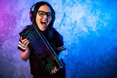 Kadın esport oyuncusu neon ışıkta oyun teçhizatıyla poz veriyor. Flamalı kız elinde klavyeyle duruyor..