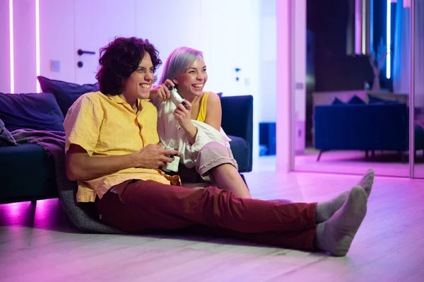 Young couple playing video games while sitting on the living room floor lit with neon color. — Stock Photo, Image