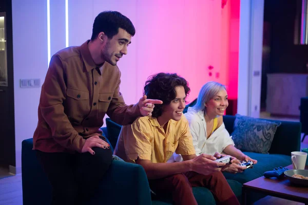 Счастливые молодые люди играют в видеоигры на консоли, сидя на диване перед телевизором. Мужчина советует своему другу в игре. Номер с теплым и неоновым освещением. — стоковое фото