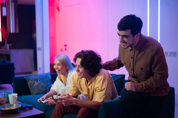 Jóvenes felices jugando videojuegos en la consola mientras están sentados en el sofá frente a la televisión. El hombre aconseja a su amigo en un juego. Habitación con luces cálidas y neón. — Foto de Stock