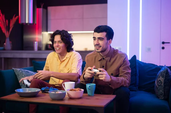 Sonriendo a diversos amigos masculinos con mandos jugando videojuegos en casa. Retrato de chicos hipster latinos y caucásicos jugando con la consola relajándose en el sofá en la sala de estar — Foto de Stock