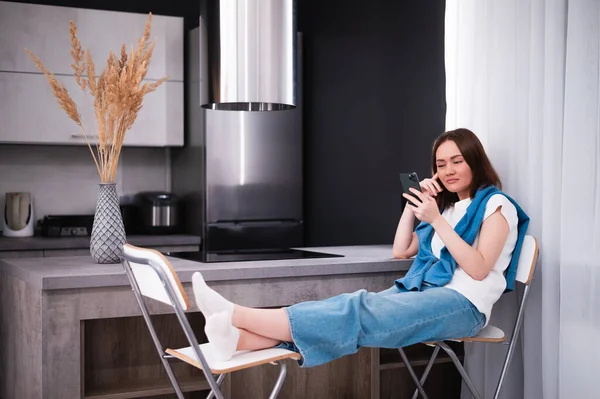 Woman bored and yawning watching media on mobile phone sitting on a chair at kitchen at home — Stock Photo, Image