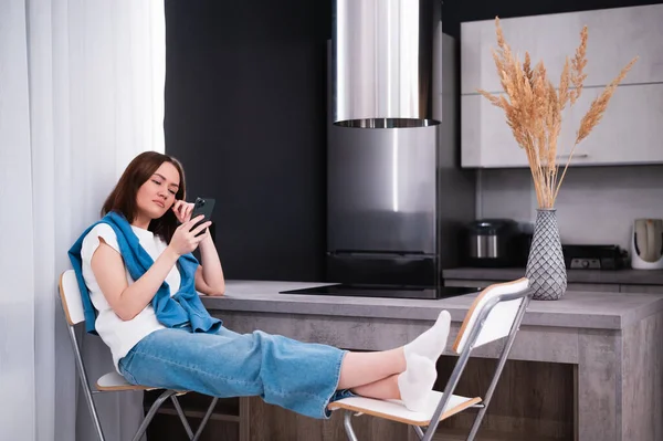 Young beautiful woman using cell phone while sitting in the kitchen after coming back home from work. — Stock Photo, Image