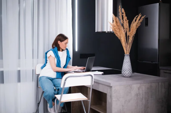 Glückliche junge schöne Frau mit Laptop arbeitet aus der Ferne, während sie zu Hause in der Küche sitzt. — Stockfoto