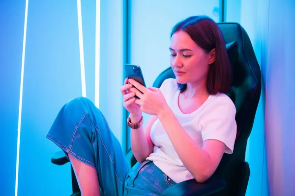 Cybersport gamer παίζει κινητό παιχνίδι στο έξυπνο τηλέφωνο κάθεται σε μια καρέκλα τυχερών παιχνιδιών σε νέον χρώμα δωματίου. — Φωτογραφία Αρχείου