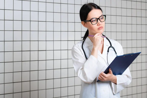 Young beautiful female doctor in white surgical coat with black stethoscope and blue paper holder in hands standing at medical office