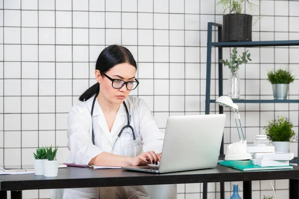 Joven mujer sonriente sentada en el escritorio, trabajando en la computadora con documentos médicos en la oficina de luz en el hospital. Doctora en bata médica, estetoscopio en consultorio. Concepto de medicina sanitaria. — Foto de Stock