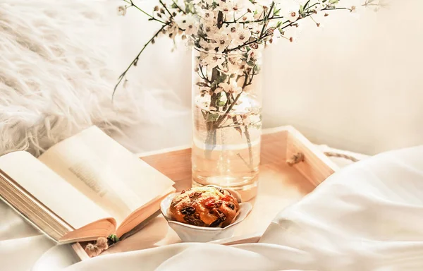 The atmosphere of a romantic morning, breakfast in bed. Flowering branches in a vase, an open book and a biscuit on a wooden tray on a white bed. Home interior. Lifestyle