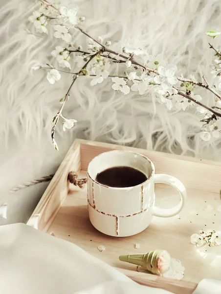 The atmosphere of a romantic morning, coffee in bed. Flowering branches in a vase, an open book and a cup of coffee on a wooden tray on a white silk bed. Home interior. Life style