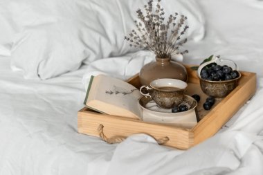 Yatakta kahve. Romantik bir sabah havası. Açık bir kitaptaki bir fincan kahve ve beyaz bir yatakta ahşap bir tepside yaban mersini. İç mekan, yaşam tarzı.