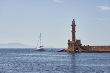 Girit 'teki Hania limanında eski bir deniz feneri ve arkasında açık mavi bir gökyüzü olan bir yat. Boşluğu kopyala.