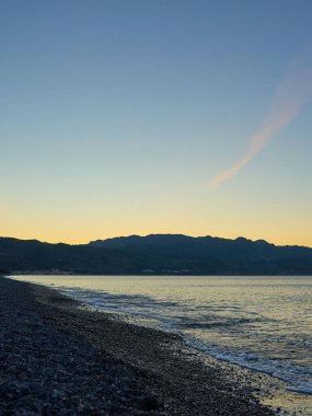 Girit 'te gün batımında çakıl taşı sahilinde bulanık dalgalar. Boşluğu kopyala.