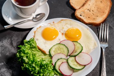 İki sahanda yumurta, marul, salatalık ve beyaz tabakta turp dilimleri, tost ve bir fincan çay veya kahve. Çırpılmış yumurta. Kahvaltıda. Sağlıklı beslenme. Yakın plan.