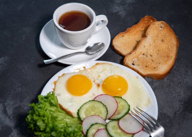 İki sahanda yumurta, marul, salatalık ve beyaz tabakta turp dilimleri, tost ve bir fincan çay veya kahve. Çırpılmış yumurta. Kahvaltıda. Sağlıklı beslenme. Yakın plan.