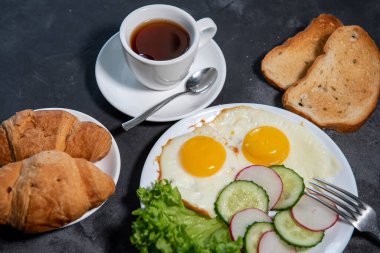 İki sahanda yumurta, bir yaprak marul, beyaz tabakta salatalık ve turp dilimleri, tost, kruvasan ve bir fincan çay veya kahve. Kahvaltıda. Sağlıklı beslenme. Yakın plan.