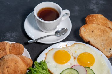 İki sahanda yumurta, bir yaprak marul, beyaz tabakta salatalık ve turp dilimleri, tost, kruvasan ve bir fincan çay veya kahve. Kahvaltıda. Sağlıklı beslenme. Yakın plan.