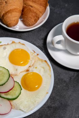 İki sahanda yumurta, bir yaprak marul, beyaz tabakta salatalık ve turp dilimleri, tost, kruvasan ve bir fincan çay veya kahve. Kahvaltıda. Sağlıklı beslenme. Yakın plan.