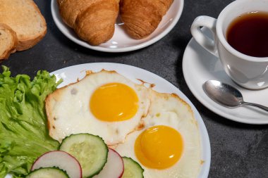 İki sahanda yumurta, bir yaprak marul, beyaz tabakta salatalık ve turp dilimleri, tost, kruvasan ve bir fincan çay veya kahve. Kahvaltıda. Sağlıklı beslenme. Yakın plan.