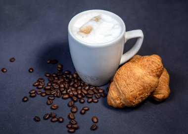 Süt köpüklü sütlü latte macchiato yanında kruvasan ve kahve çekirdeği ile birlikte. Koyu mavi arka plan. Bir fincan sütlü kahve. Süt üzerine fotokopi alanı olan sıcak latte ya da kapuçino.