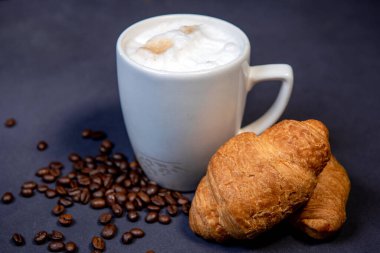 Süt köpüklü sütlü latte macchiato yanında kruvasan ve kahve çekirdeği ile birlikte. Koyu mavi arka plan. Bir fincan sütlü kahve. Süt üzerine fotokopi alanı olan sıcak latte ya da kapuçino.