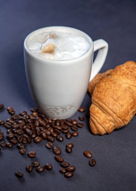 Süt köpüklü sütlü latte macchiato yanında kruvasan ve kahve çekirdeği ile birlikte. Koyu mavi arka plan. Bir fincan sütlü kahve. Süt üzerine fotokopi alanı olan sıcak latte ya da kapuçino.