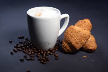 Süt köpüklü sütlü latte macchiato yanında kruvasan ve kahve çekirdeği ile birlikte. Koyu mavi arka plan. Bir fincan sütlü kahve. Süt üzerine fotokopi alanı olan sıcak latte ya da kapuçino.