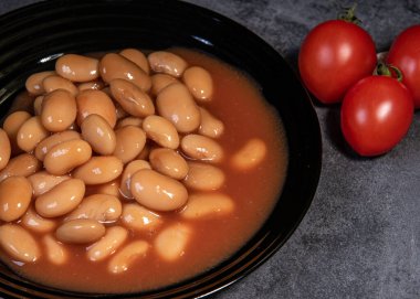 Domates soslu beyaz falo. Kaynamış fasulye, siyah bir tabakta domates sosu ve vişneli domates. Yakın plan.