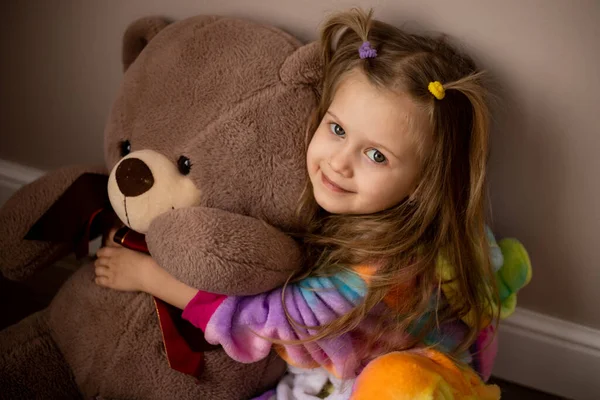 Çok renkli kegurumisiyle at kuyruklu küçük güzel bir kız büyük bir oyuncak ayıya sarılıyor ve kameraya bakıyor.
