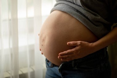 hamile bir kadın pencerenin önünde, üçüncü üç aylık dönemde çıplak karnına dokunup okşadı. Çocuk beklentisi, doğum hazırlığı, hamilelik ve annelik kavramı. Seçici odak