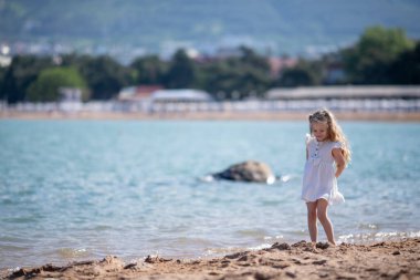 Uzun saçlı, beyaz elbiseli, kumsalda kumla oynayan tatlı bir kız. Dağların ve Karadeniz 'in güzel manzarası, Gelendzhik tatil köyü. Seçici odak