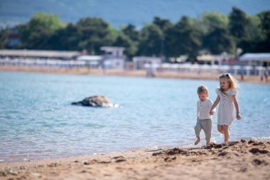 Küçük oğlan ve kız yazın deniz kıyısında el ele tutuşarak koşarlar. Erkek ve kız kardeşlerin doğal kıyafetleri. Turizm, tatiller, çocuklarla seyahat, güneş koruma ve güvenlik
