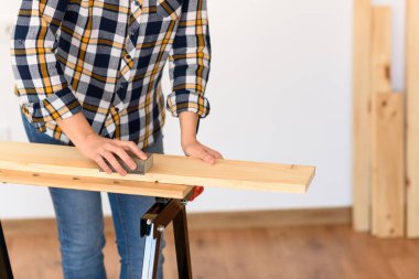 Ekose gömlek ve kot pantolon giymiş, el işi yapan bir kız. Beyaz tenli ve kıvırcık saçlı. Beyaz bir arka planı ve ahşap tahtaları olan bir tezgahın üzerine zımparalıyor..