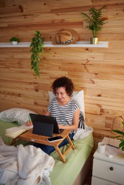 A curly-haired woman teleworks from her bed in concentration. The room has a nice wooden wall and it is daytime. New technologies allow you to work comfortably. She uses a folding table. clipart