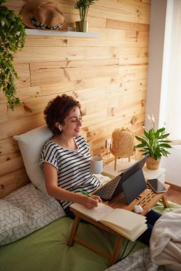 A curly-haired woman drinks coffee while working from her bedroom. The room has a nice wooden wall and it is daytime. New technologies allow you to work comfortably. She uses a folding table. clipart