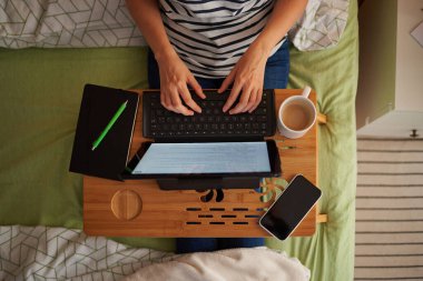 Top view of a woman telecommuting from her bed. He types on her laptop, with a mug and notebook on the table. The room has a decorative wooden wall. clipart