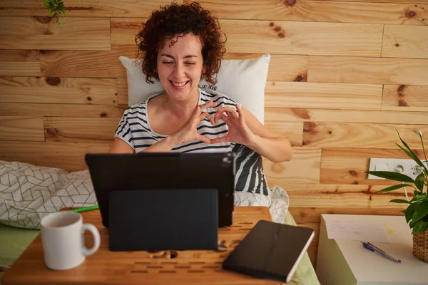 A curly-haired Caucasian woman draws a heart with her hands while talking to her loved ones through a video call. She uses a folding table and a tablet. She is leaning against a wooden wall.
