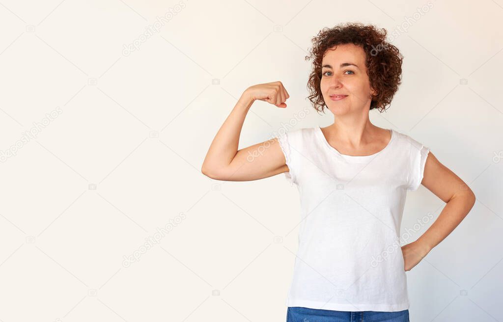 A curly haired Caucasian young woman shows off her bicep muscles as a sign of success and health. White background. Isolated person.