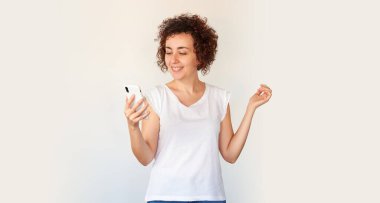 Akıllı telefona bakan, cep telefonu çığlığı atan ve beyaz stüdyo arka planında izole edilmiş sevinçle cep telefonu çığlığı atan inanılmaz alışveriş mobil uygulaması satış mesajına hayran olan genç bir kadın..