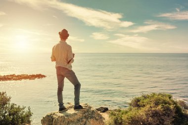 Yüzü görünmeyen genç bir gezgin, şapka, gömlek ve kot giyiyor, güneş batarken turistik bir plaj bölgesinde kamera tutuyor.