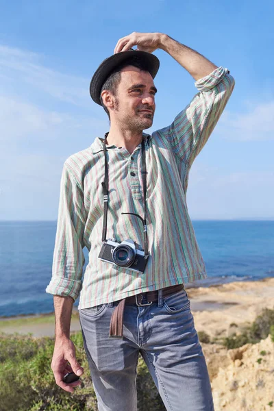 A young tourist man standing in front of the camera with the sea in the background happy to be able to travel and discover places with his photography equipment
