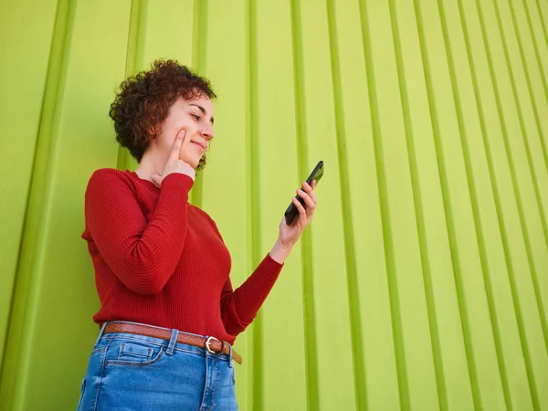 A young Caucasian woman with curly hair wearing a red sweater and jeans uses her mobile phone to surf the internet, she has a green wall in the background and it is daytime