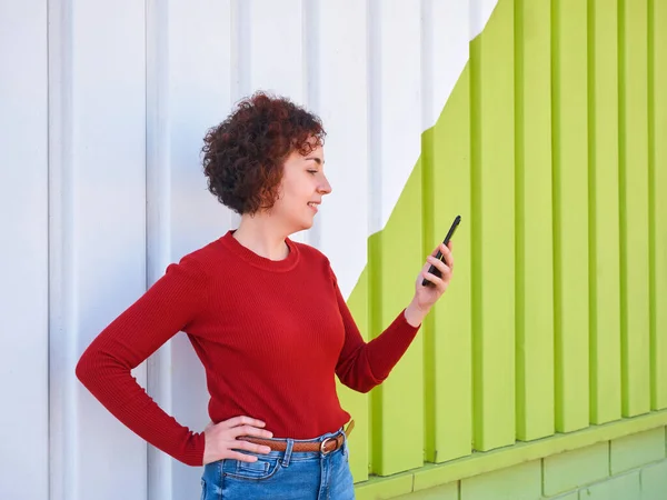 A young Caucasian woman with curly hair wears a red sweater and jeans, is looking at the screen of her mobile phone in front of a white and green wall, it is day and there is natural light