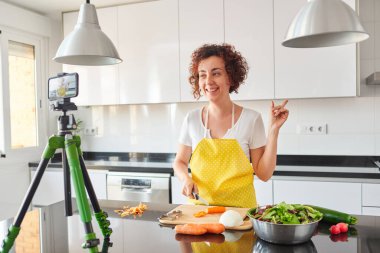 Kadın kendini evinin mutfağında kamerayla kaydediyor. Salata tarifi hazırlarken doğal ışık var. Sarı önlük ve beyaz tişört giyiyor.