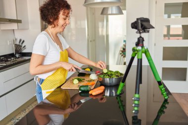 Kadın kendini evinin mutfağında kamerayla kaydediyor. Salata tarifi hazırlarken doğal ışık var. Sarı önlük ve beyaz tişört giyiyor.