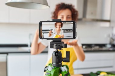 Kadın kendini akıllı telefonuyla mutfağında salata tarifi hazırlarken kaydediyor. Doğal ışık var ve sarı önlük ve beyaz tişört giyiyor. Kafkasyalı.