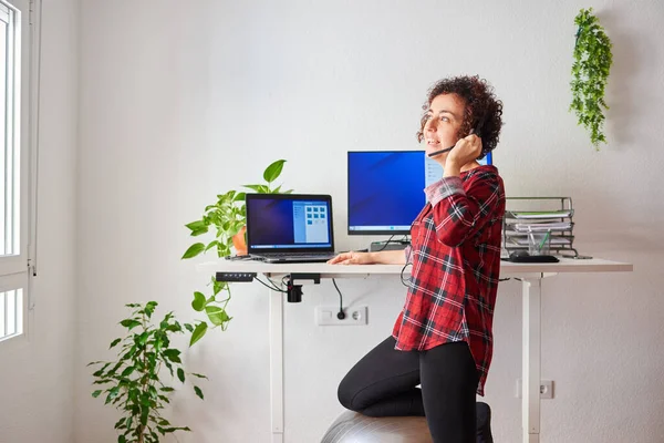 Woman teleworking at an adjustable standing desk with one knee resting on a fitball