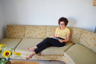 Genç kadın kanepeye oturmuş dijital tabletine resim çiziyor.