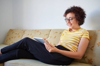 Genç kadın kanepeye oturmuş dijital tabletine resim çiziyor.