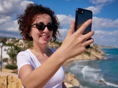 Arka planda deniz olan cep telefonuyla selfie çeken bir kadın.
