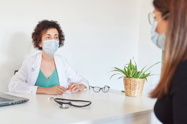 Yüz maskeli bir kadın doktor ofisinde bir hastayla ilgileniyor.